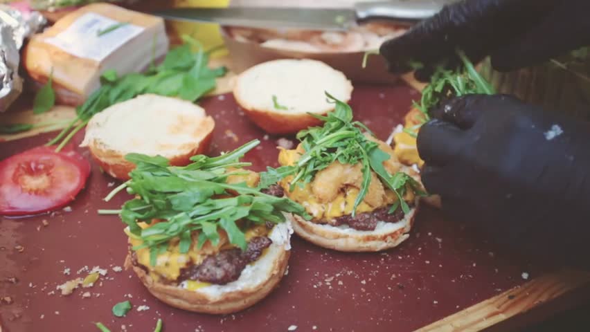 A vendor prepares fresh, flavorful hamburgers topped with greens, cheese, and sauce at a lively street food festival, showcasing the art of street-style gourmet cooking.
