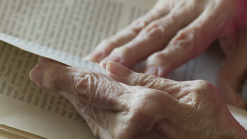Elderly woman's hands, deformed by arthritis, turn the pages of an old book.