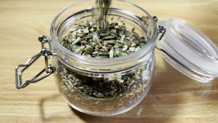Closeup slow motion shot of mixed edible seeds (sunflower, pumpkin, hemp and linseed) pouring into an open glass storage jar, with an airtight lid and metal clasp.