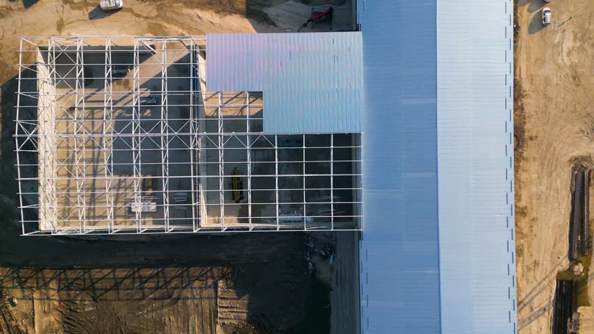 A construction site with a partially built warehouse featuring a steel framework and metal roof. Workers and heavy machinery operate on-site, showcasing industrial progress and structural development