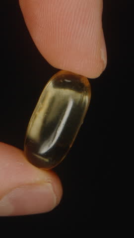 Vertical video. A Hand Holding a Single Golden Transparent Omega 3 Capsule Highlighting Its Smooth Texture and Glossy Finish Against a Black Background Nutritional Supplementation