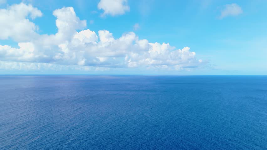 Low fast aerial dolly rises above blue ocean water with soft ripples under a bright sky, serene nature backdrop