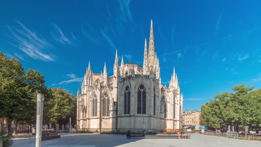 Cathedrale Saint-Andre de Bordeaux timelapse hyperlapse with twin spires under a vibrant blue sky. Green trees and people walking in the square create a serene urban atmosphere. Bordeaux, France