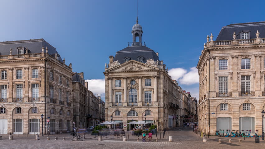 Place de la Bourse timelapse hyperlapse in Bordeaux, France, with restaurants surrounded by classic architecture under a cloudy blue sky. People walking around the main square and central streets