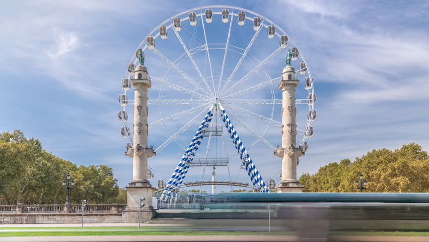 Large Ferris wheel between two columns at Place des Quinconces during the Foire aux Plaisirs timelapse in Bordeaux, France. Bright blue sky, white clouds and vibrant atmosphere. Traffic on the street