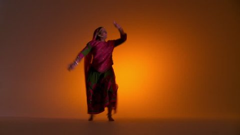 Indian girl in traditional folk costume dances energetically an orange colored background with flashing lighting. Medium close up.