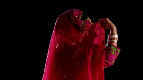 Indian girl in traditional dance costume looks to the camera, then looks away, covering her face with her veil. Recorded against black background.