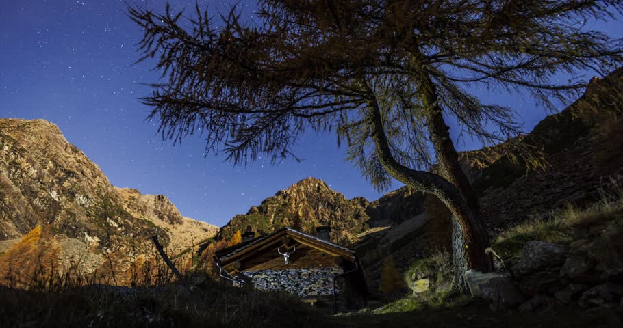 Enchanting timelapse video capturing star trails over a rustic cabin and autumnal tree in Predaccio, surrounded by majestic mountain peaks under a vivid night sky