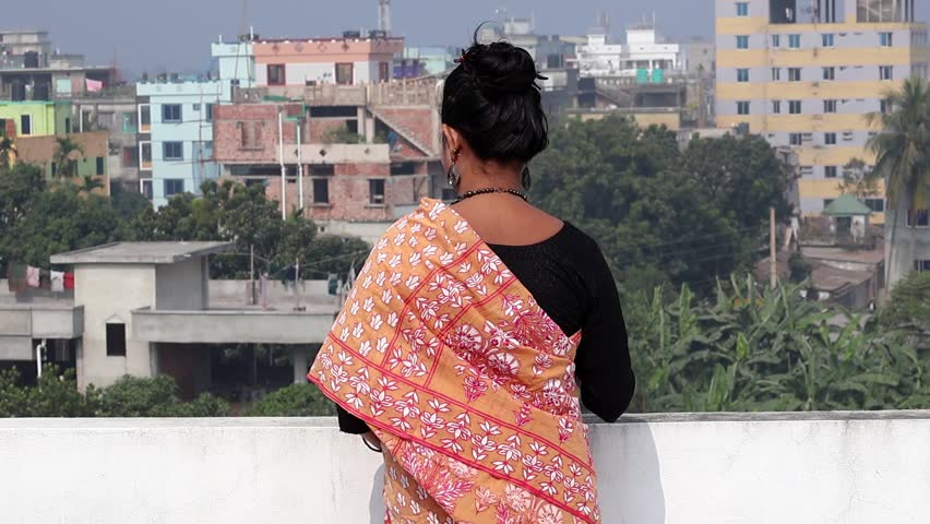 A Bengali woman, draped in a saree, stands alone on a rooftop, gazing into the distance, lost in thought. Slow-motion video footage. Back view.