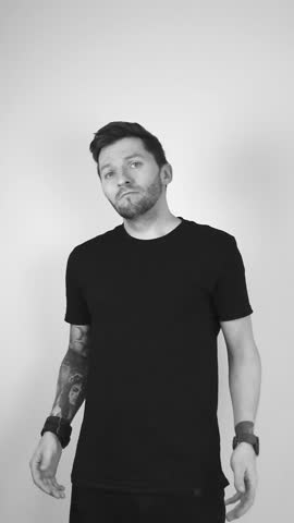Grayscale footage of a white tattooed man with short hair and beard raising his arms in confusion showing that he does not know anything or is oblivious. Raising both arms, palms up, tilting his head.
