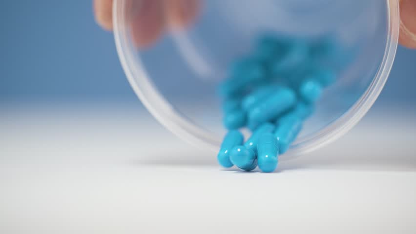 A cascade of blue pills spills onto a clean white surface in slow motion 4K. The vibrant color and glossy textures create a visually dynamic and polished aesthetic, with scattered motion