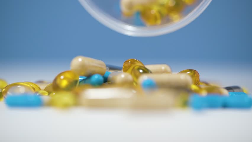 An array of colorful tablets and capsules spills dramatically onto a white surface in slow motion 4K. The vibrant blue background emphasizes the chaotic movement, creating a stunning