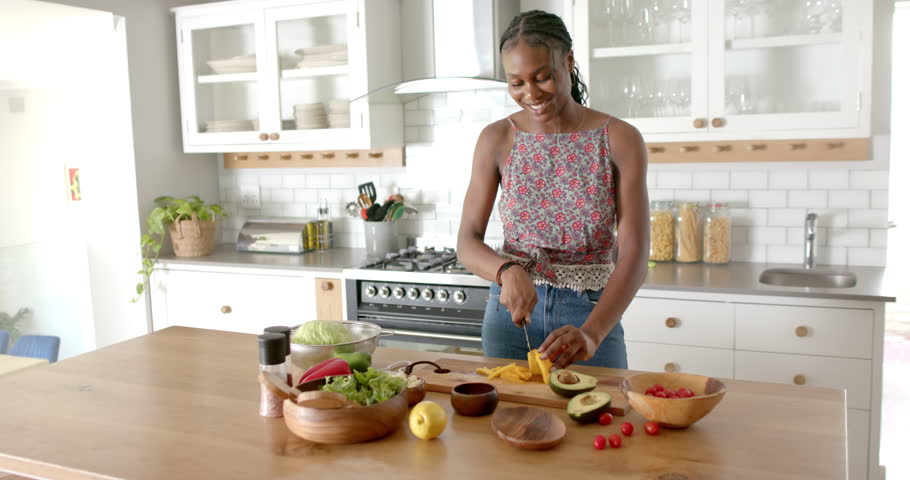 African American young woman wearing floral top is cutting vegetables in a kitchen, at home. She has braided hair, a bright smile, and is preparing a salad, unaltered, slow motion