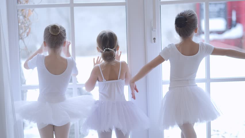 Three young girls in white ballet dresses are standing in front of a window, watching the snow fall outside