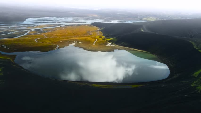 Aerial drone footage of a large lake in a crater that has sharp steep mountain cliff edges covered in green moss and plants. River has brown plants and grass around in Greenland or Iceland, cloudy day