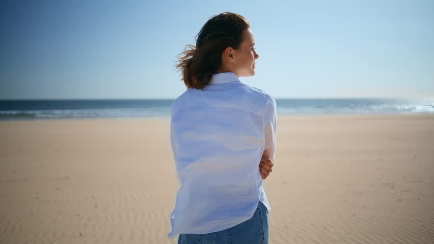 Back view woman walking beach in white shirt beautiful windy day. Young brunette looking ocean horizon enjoying summer vacation on seashore. Serene lady strolling gold coastal sand feeling freedom.
