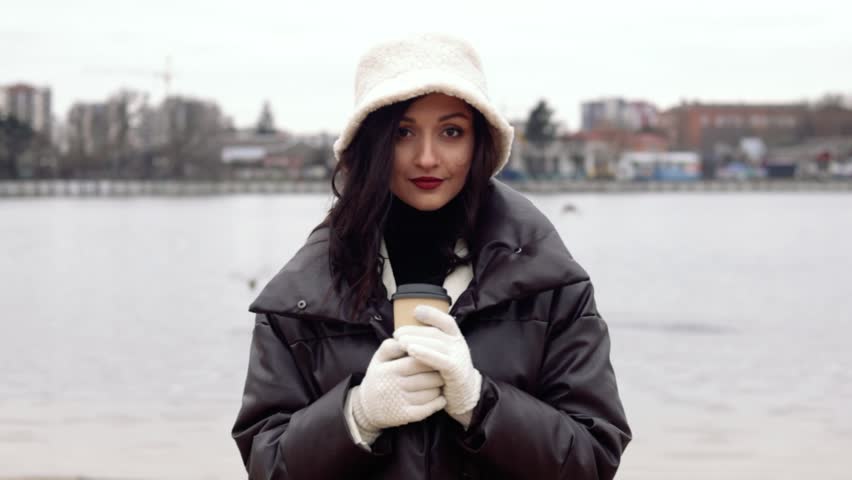 Fashionable young woman wearing a winter coat, gloves, and a bucket hat, holding a coffee cup near a river on a cold day