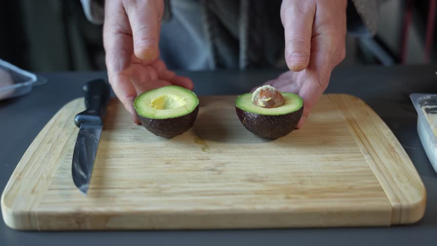 A person is cutting an avocado in half on a wooden cutting board. The knife is on the left side of the avocado