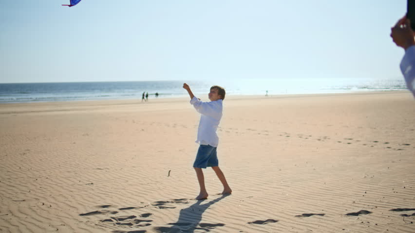 Boy flying colorful kite on sandy beach. Happy mother filming playful child running energetically near ocean in bright summer sunlight. Active family spending weekend leisure together at seashore.