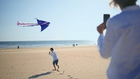 Boy flying colorful kite on sandy beach. Happy mother filming playful child running energetically near ocean in bright summer sunlight. Active family spending weekend leisure together at seashore. - Powered by Shutterstock - Get 15% off with code: PIKWIZARD15