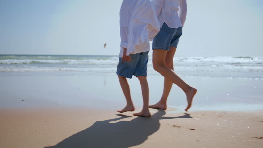 Mother boy walking barefoot on wet sand by sea waves closeup. Unknown woman strolling with beloved son on sunny seashore bonding. Happy family spending time at shoreline enjoying relaxed connection.