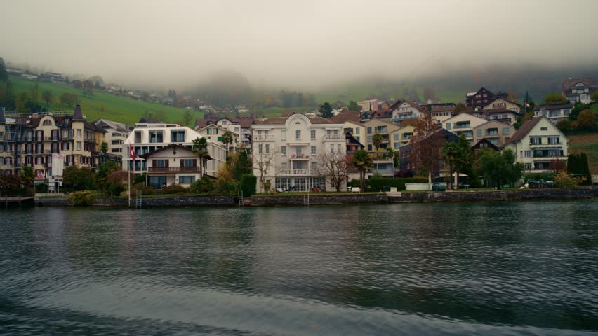Colorful lakeside houses and villas surrounded by lush greenery on a foggy day near Lucerne, Switzerland. The serene water reflects the scenic architecture and landscape.