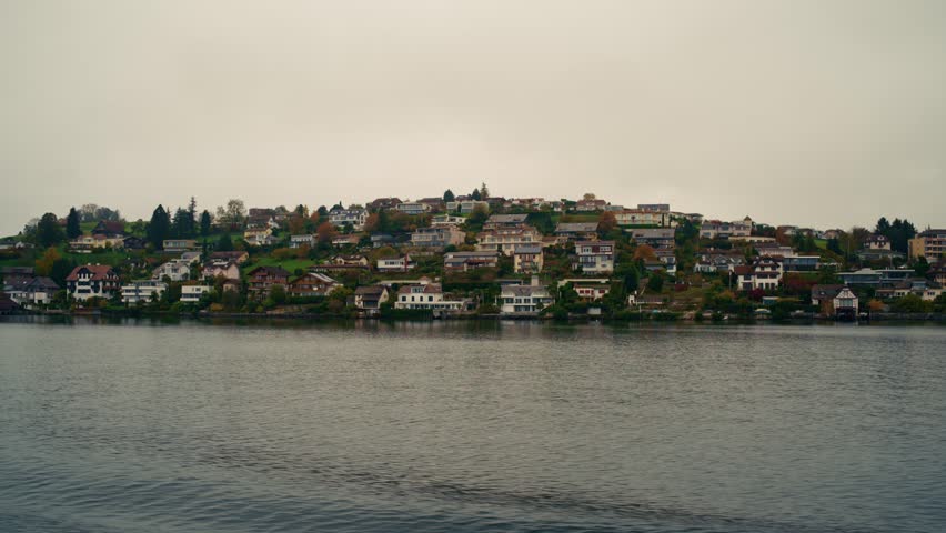 Scenic view of a hillside neighborhood in Lucerne, Switzerland, with traditional Swiss houses and lush greenery reflecting on the calm lake under a cloudy sky.