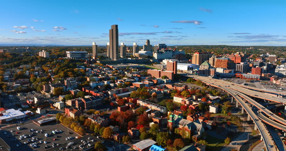 Approaching a high-rise building standing out from the downtown scenery. Aerial view of the vast green cityscape of Albany, New York State, USA on sunny day.