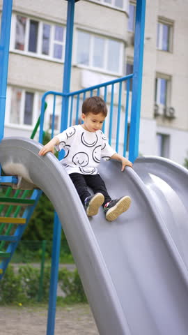 Beautiful kid goes down the slide and smiles to the camera. Cheerful baby boy plays on the playground in summer. Vertical video.