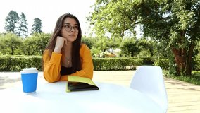 Young brunette woman with glasses wearing orange shirt sitting at table against nature background. Woman writing something in notebook with pen in botanical garden - Powered by Shutterstock - Get 15% off with code: PIKWIZARD15
