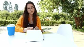 Young brunette woman with glasses wearing orange shirt sitting at table against nature background. Woman writing something in notebook with pen in botanical garden - Powered by Shutterstock - Get 15% off with code: PIKWIZARD15