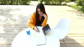 Young brunette woman with glasses wearing orange shirt sitting at table against nature background. Woman writing something in notebook with pen in botanical garden - Powered by Shutterstock - Get 15% off with code: PIKWIZARD15