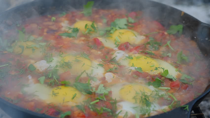 Recipe and cooking process for Shakshuka at winter time. Close up of male hand puts green parsley in scrambled egg in frying pan with chopped bell peppers, tomatoes and onions
