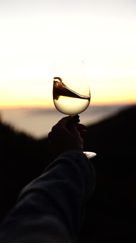 Super slow motion close-up of white wine swirling in a transparent glass in the hands of a young girl against the backdrop of a sunset for taste testing and quality control. Vertical video.