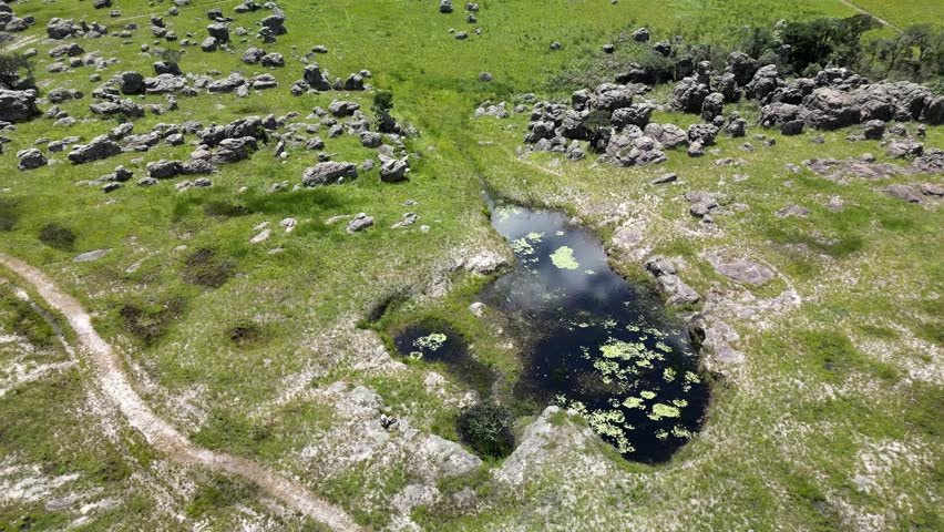 A high aerial reverse of a woman relaxing at a pond in nature, surrounded by lush green grassland, metamorphic rock formations, and rolling green hills in Africa.