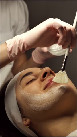 A beautician applies a white cleansing mask to a female face close-up, vertically. A woman at a relaxing spa treatment enjoys