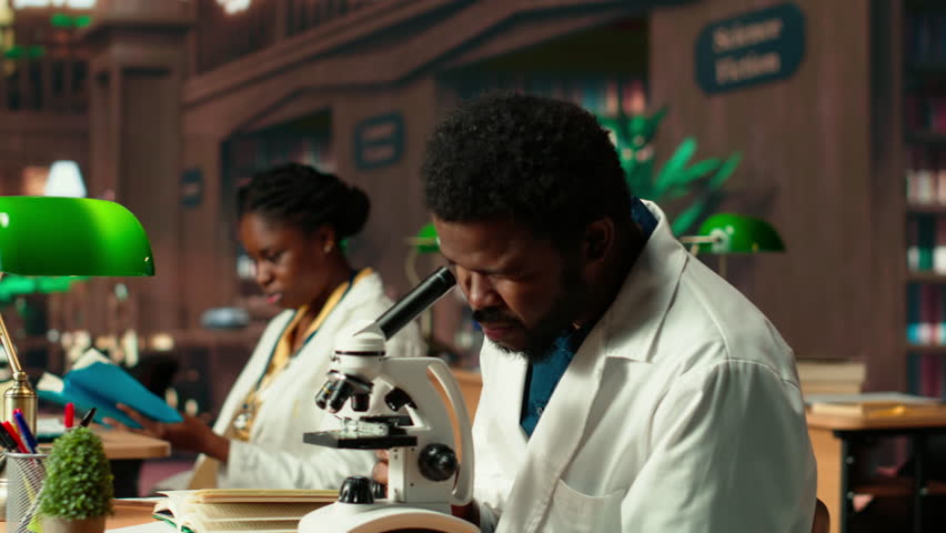 Male student researching viruses during a microscope analysis and academic reading in a classic library. Med school pupil reflects dedication to biology and exam preparations. Camera B.