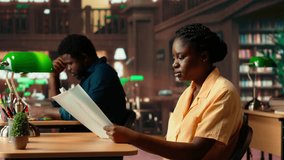 Focused african american student sits in a quiet library reading papers and class notes, studying under the warm glow of a green lamp. Woman surrounded by books and learning tools. Camera B. - Powered by Shutterstock - Get 15% off with code: PIKWIZARD15