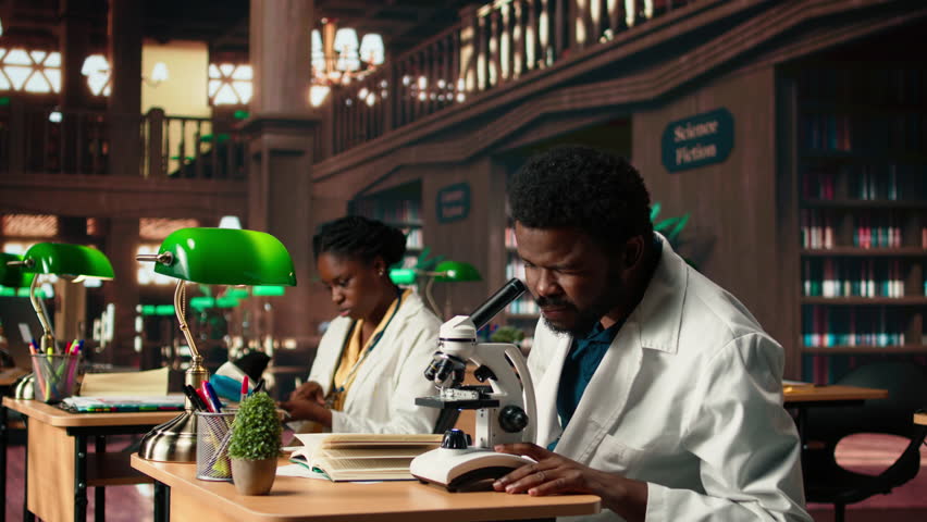 African American pupil studies microbiology in a campus library, examining cells and microorganisms under the microscope. Male student prepares for his biotechnology thesis. Camera A.