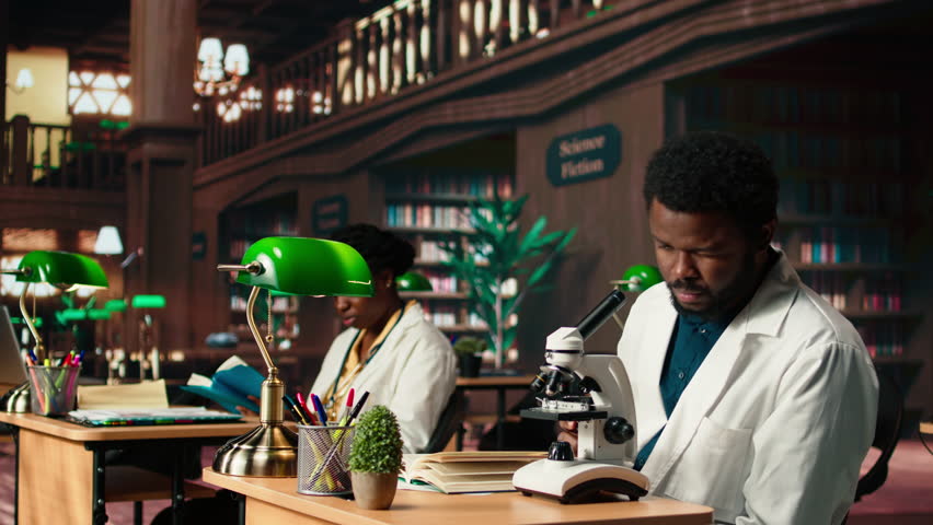 Male student researching viruses during a microscope analysis and academic reading in a classic library. Med school pupil reflects dedication to biology and exam preparations. Camera A.