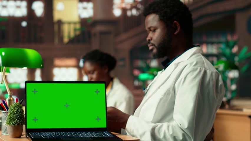Medical student uses books in the campus study space next to green screen, combining research, anatomy lessons and academic methods. Pupil reading research papers for medical practice. Camera B.