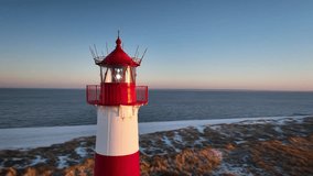 Aerial orbit of red and white striped lighthouse situated on a coastal landscape at sunrise in winter. Lighthouse West-List on the Ellenbogen (the "Elbow"), the peninsula on northern coast of Sylt isl - Powered by Shutterstock - Get 15% off with code: PIKWIZARD15