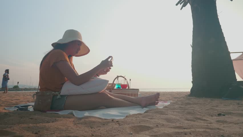 A person sits on the sandy beach, enjoying a sunny day while using a smartphone. Palm trees and a serene atmosphere create a perfect setting for relaxation and leisure by the sea.