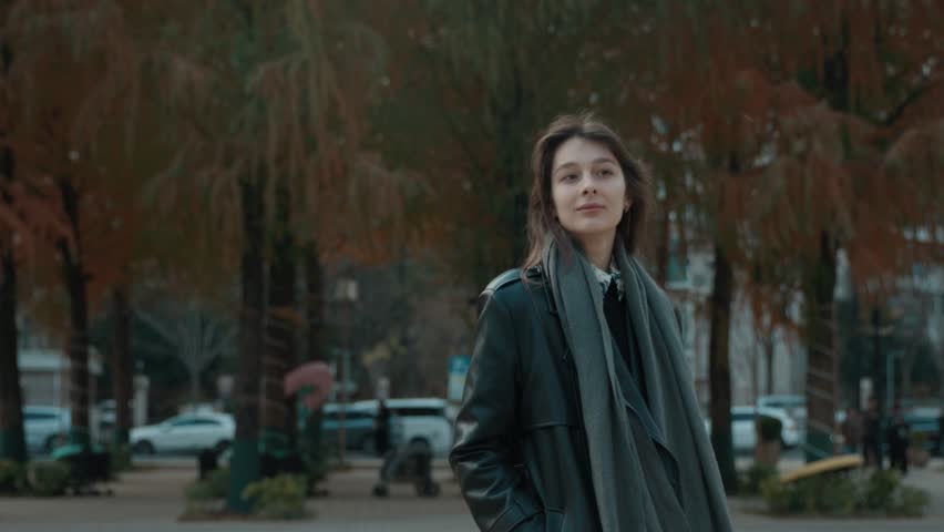A woman wearing a black coat and scarf stands in front of a tree. The scene is set in a city with cars and people around her. The woman is smiling, giving off a positive and friendly vibe