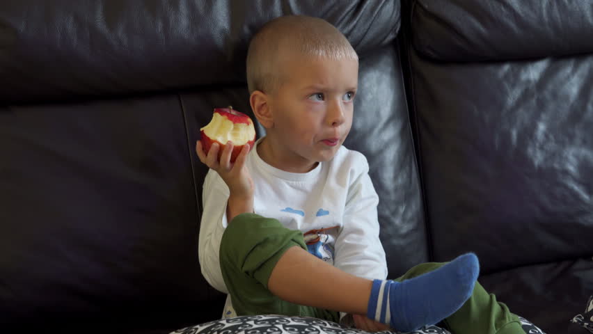 Child eating a red juicy apple with appetite. Little boy biting an apple while sitting on the couch and watching TV.