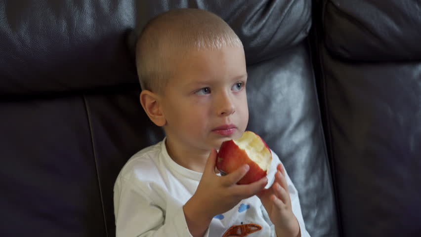 Child eating a red juicy apple with appetite. Little boy biting an apple while sitting on the couch and watching TV.