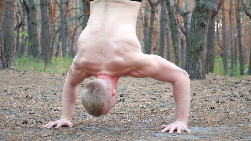 Young muscular man doing push upsin handstand at forest. Hardy and strong guy doing stunts at wood. Close up torso of athlete showing gymnastic exercises outdoor. Sportive and healthy lifestyle
