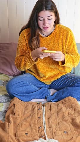 Caucasian girl with long hair sitting on her bed taking detailed video of a jacket to sell on social media. Selling second hand clothes and sustainability.