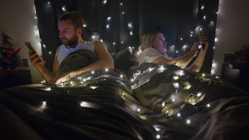 A couple relaxes in bed, each engrossed in their smartphones. The cozy atmosphere is enhanced by soft fairy lights and small festive decorations, creating a warm setting for the evening.