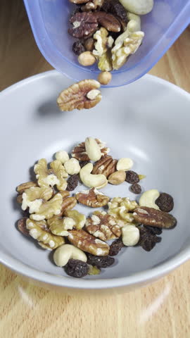 Vertical video - Closeup slow motion shot of mixed shelled nuts and raisins being poured from a container into a bowl, to eat.